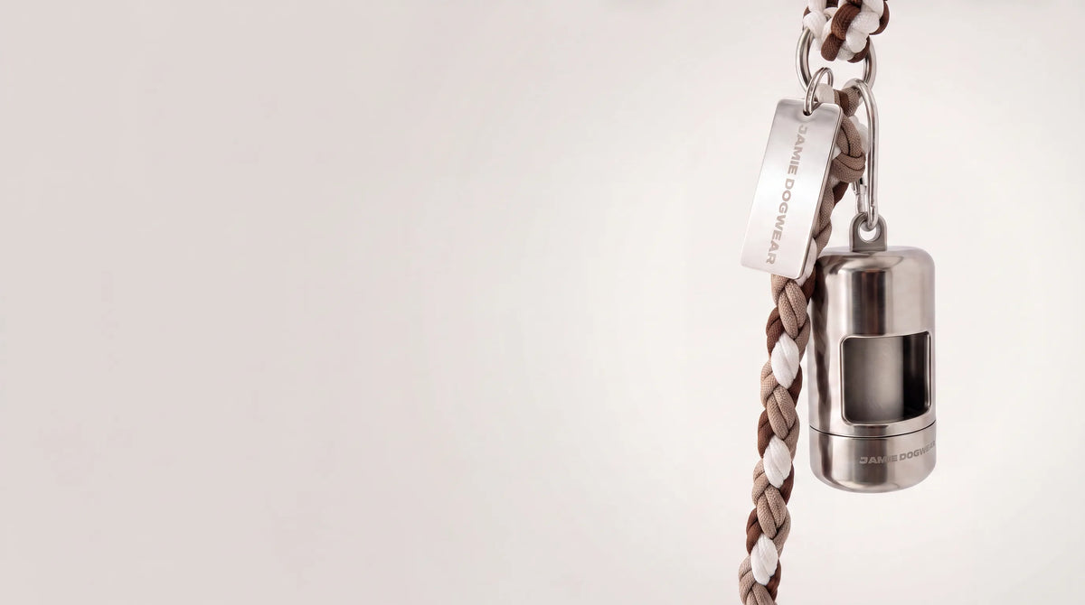 A silver dog poop bag holder attached to a brown, beige, and white braided cord leash, featuring a silver metal tag engraved with 'JAMIE DOGWEAR'.