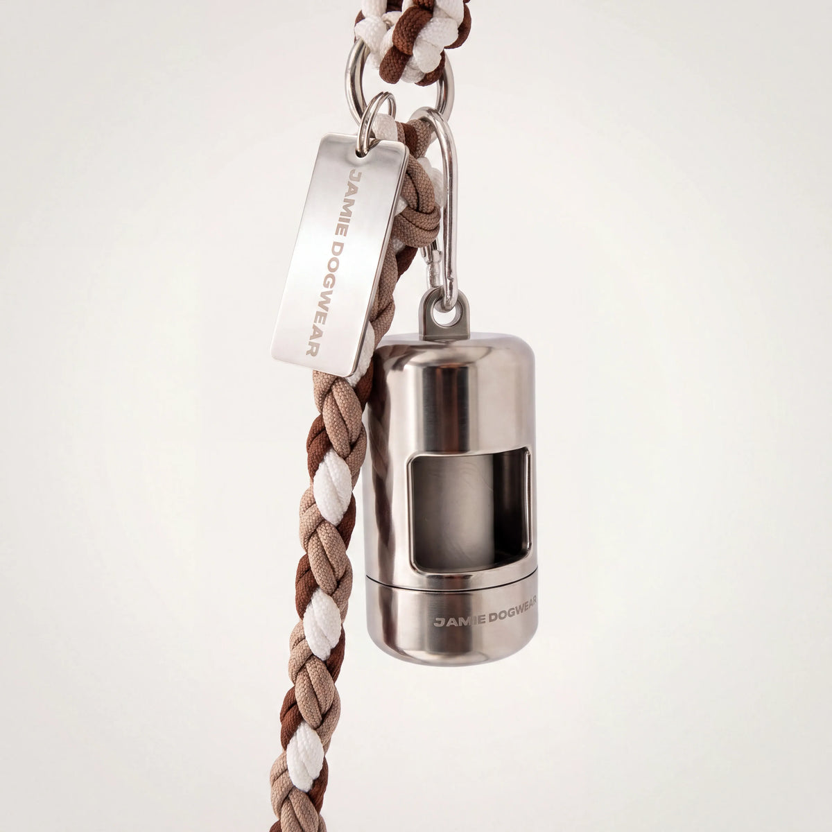 A silver dog poop bag holder attached to a brown, beige, and white braided cord leash, featuring a silver metal tag engraved with 'JAMIE DOGWEAR'.