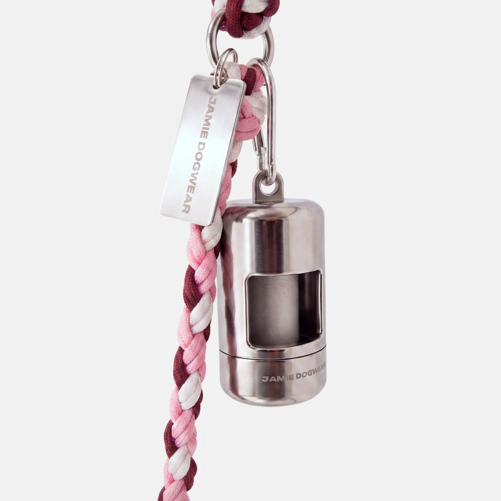 A silver dog poop bag holder attached to a pink, burgundy, and white braided cord leash, featuring a silver metal tag engraved with 'JAMIE DOGWEAR'.