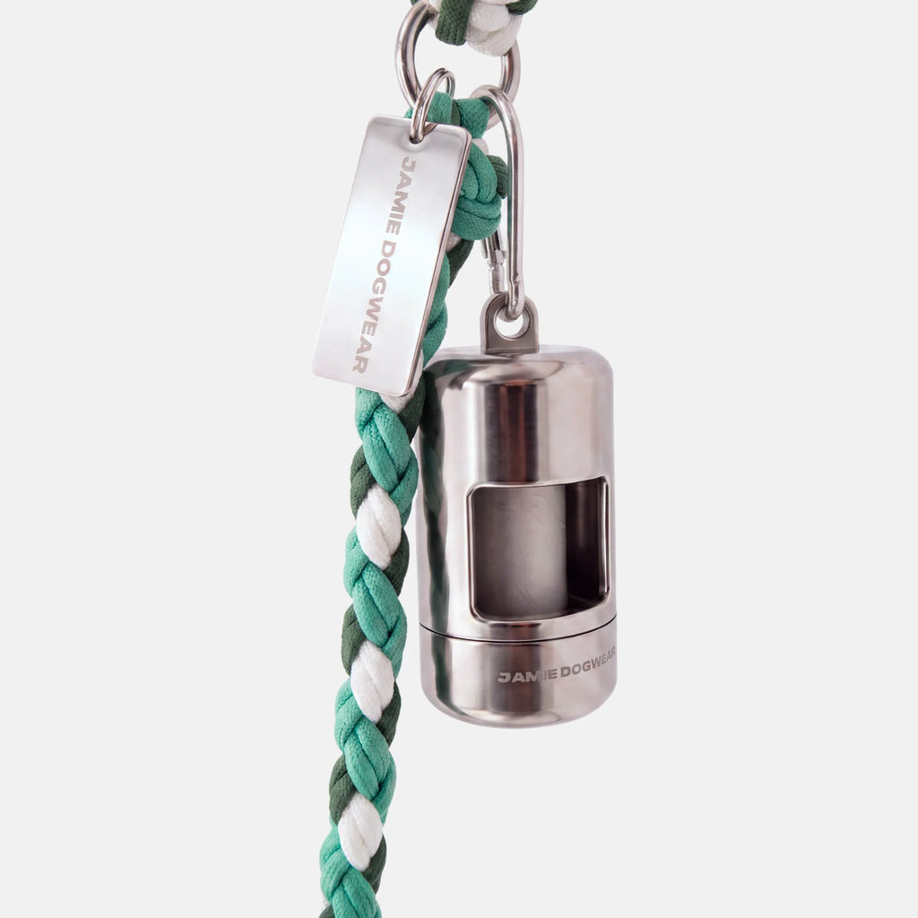 A silver dog poop bag holder attached to a green and white braided cord leash, featuring a silver metal tag engraved with 'JAMIE DOGWEAR'.