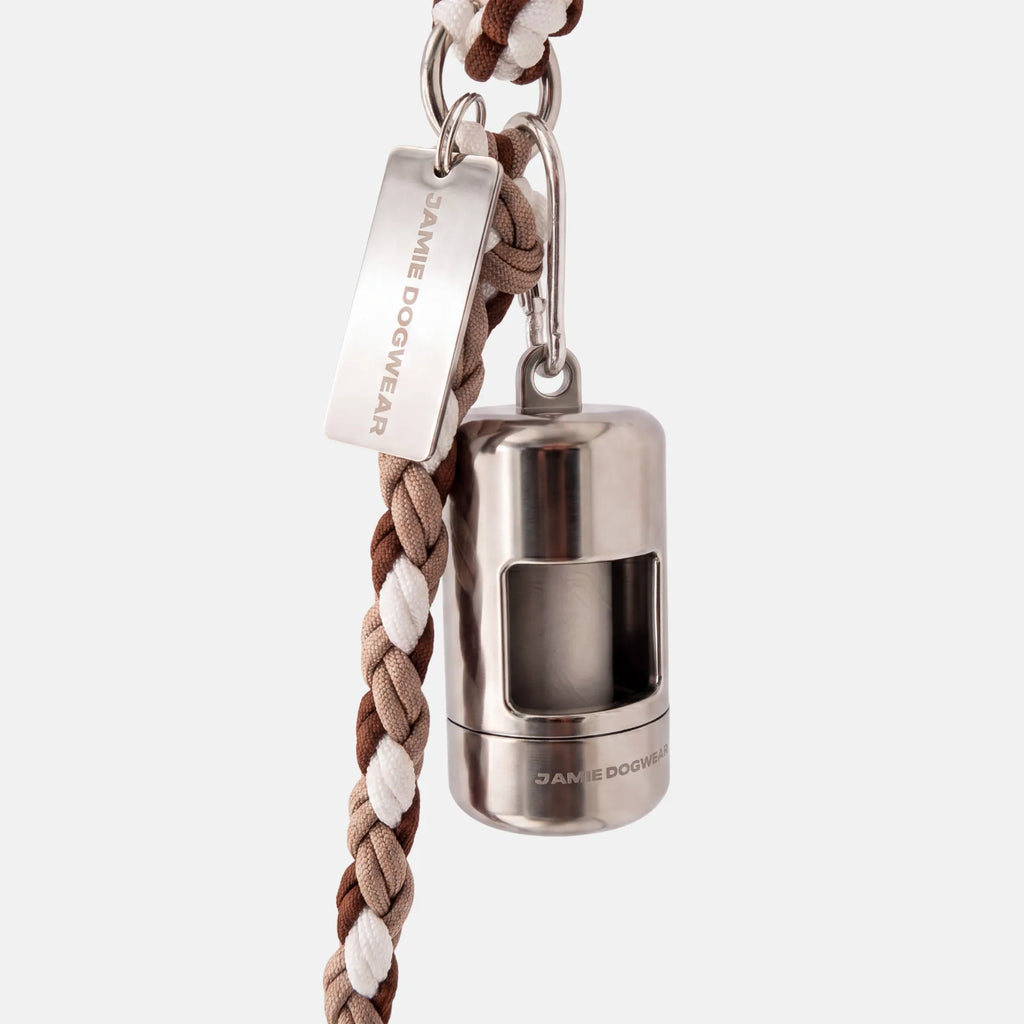A silver dog poop bag holder attached to a brown, beige, and white braided cord leash, featuring a silver metal tag engraved with 'JAMIE DOGWEAR'.